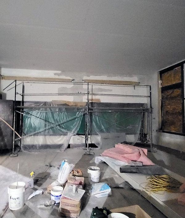 a large room is under construction with a window filled with chipboard, scaffolding, and plastic; buckets, pink insulation bats, and other construction materials are in the middle of the room