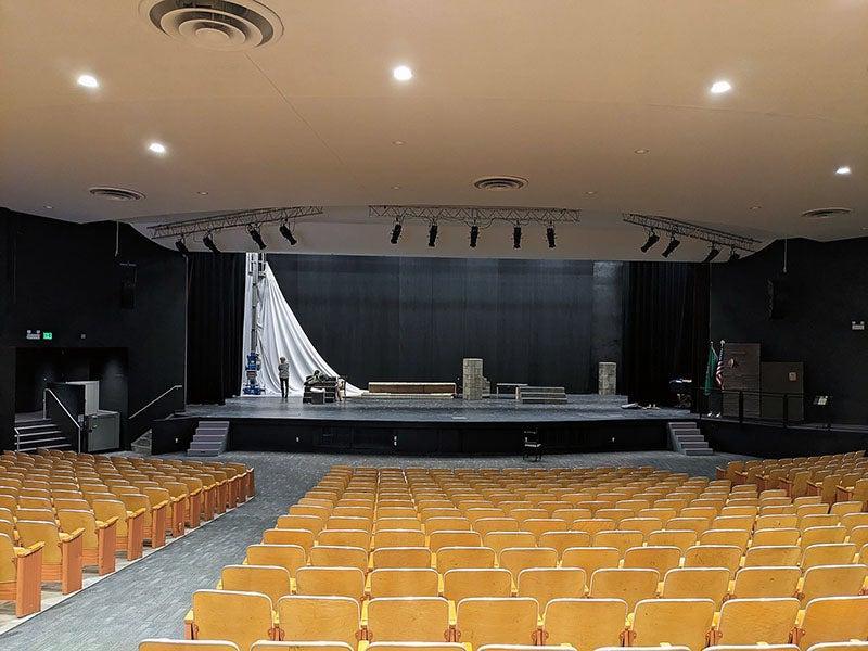 a view from behind theater seating toward a stage with a white curtain being installed at the the top