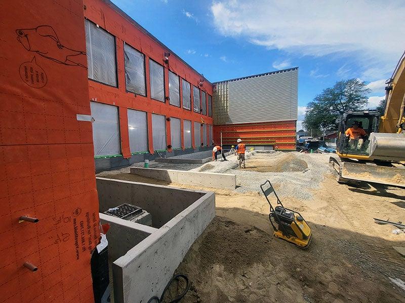 a courtyard area has soil, gravel, and concrete walls. workers are moving materials. a compactor for gravel is in the front
