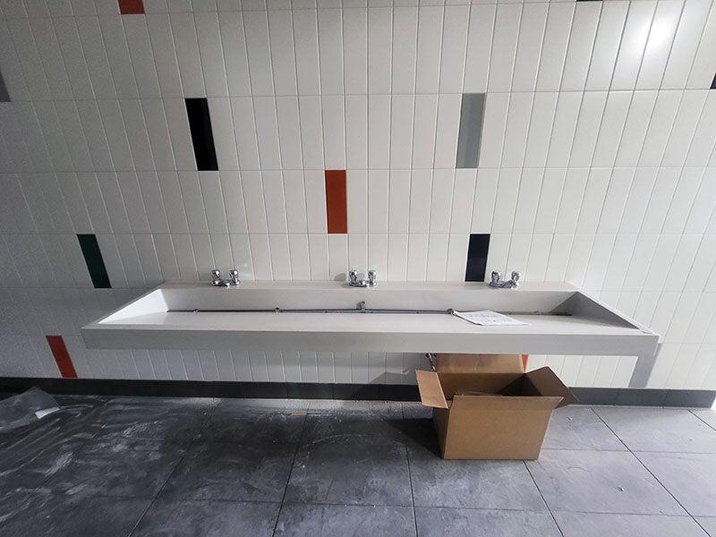 a trough sink with three faucets hangs on a tile wall with a few colored accent tiles