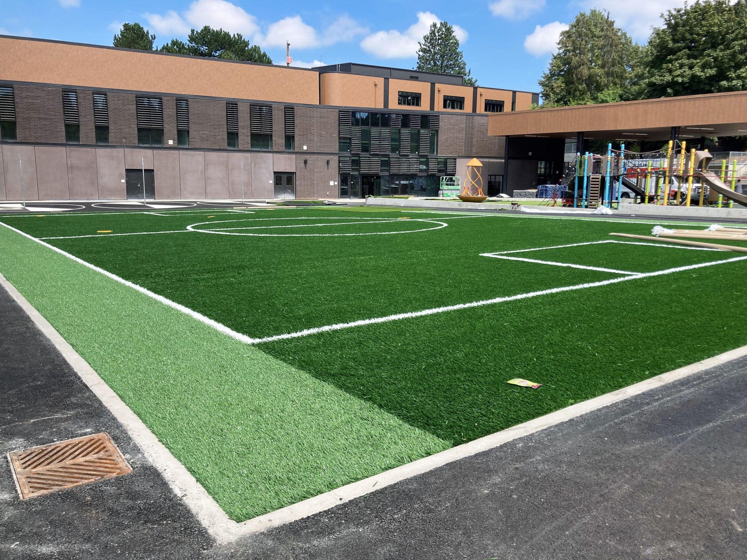 an green turf area with soccer markings is in front of a building with playground equipment behind it
