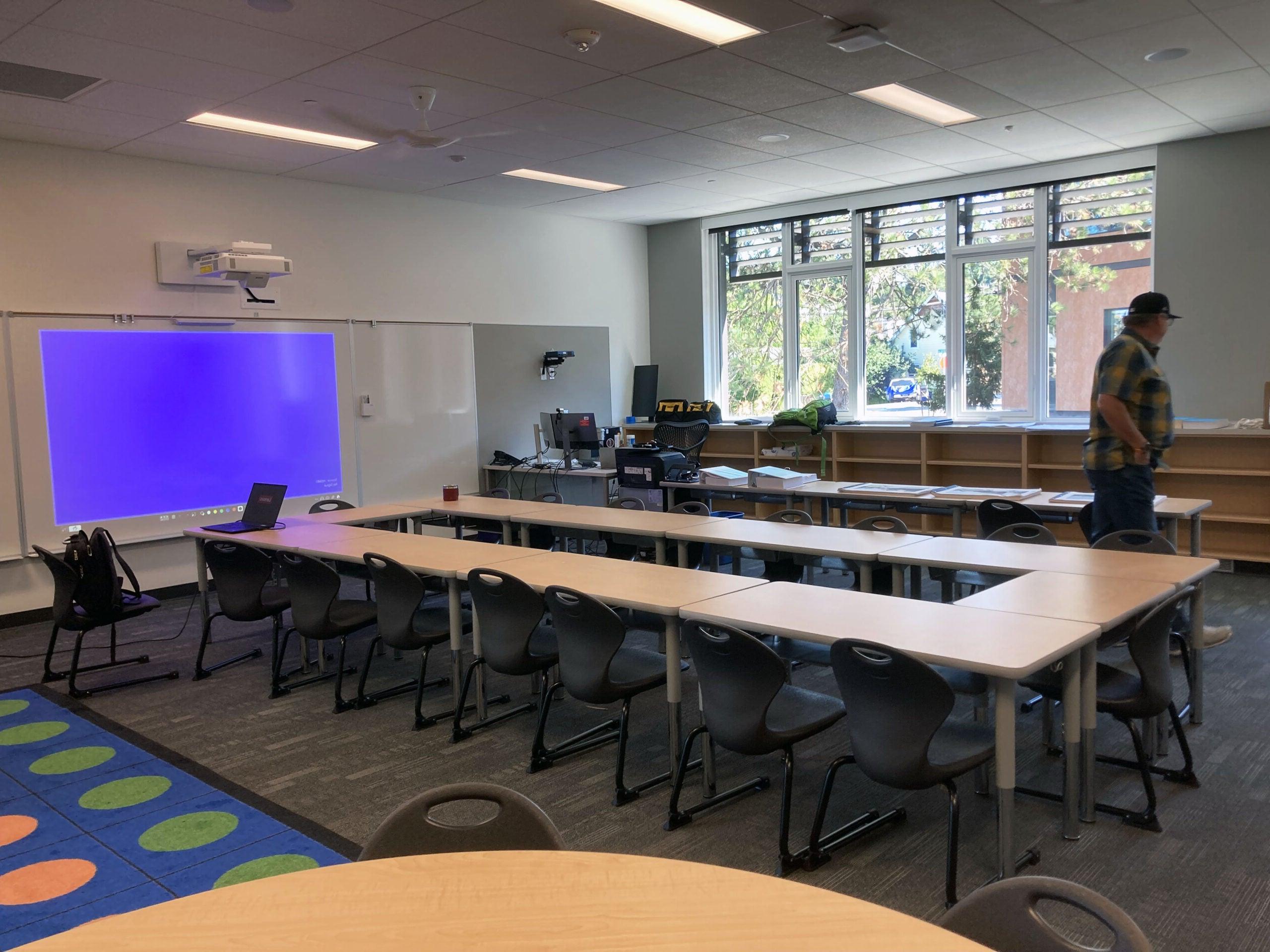 a classroom with tables set in a circle, windows on one wall with bookshelves below, a white board and projector on another wall