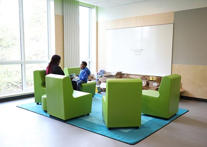 a group of lime green chairs sites on a turquoise rug in a corner with windows on both sides. an adult and a child site in the two of the seats