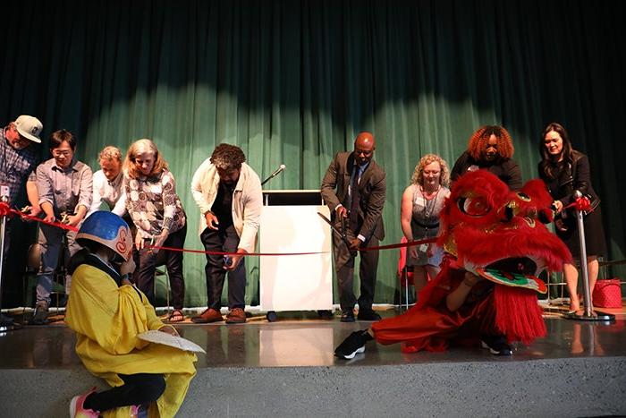 a group of people on a stage lean toward a red ribbon with scissors in their hands