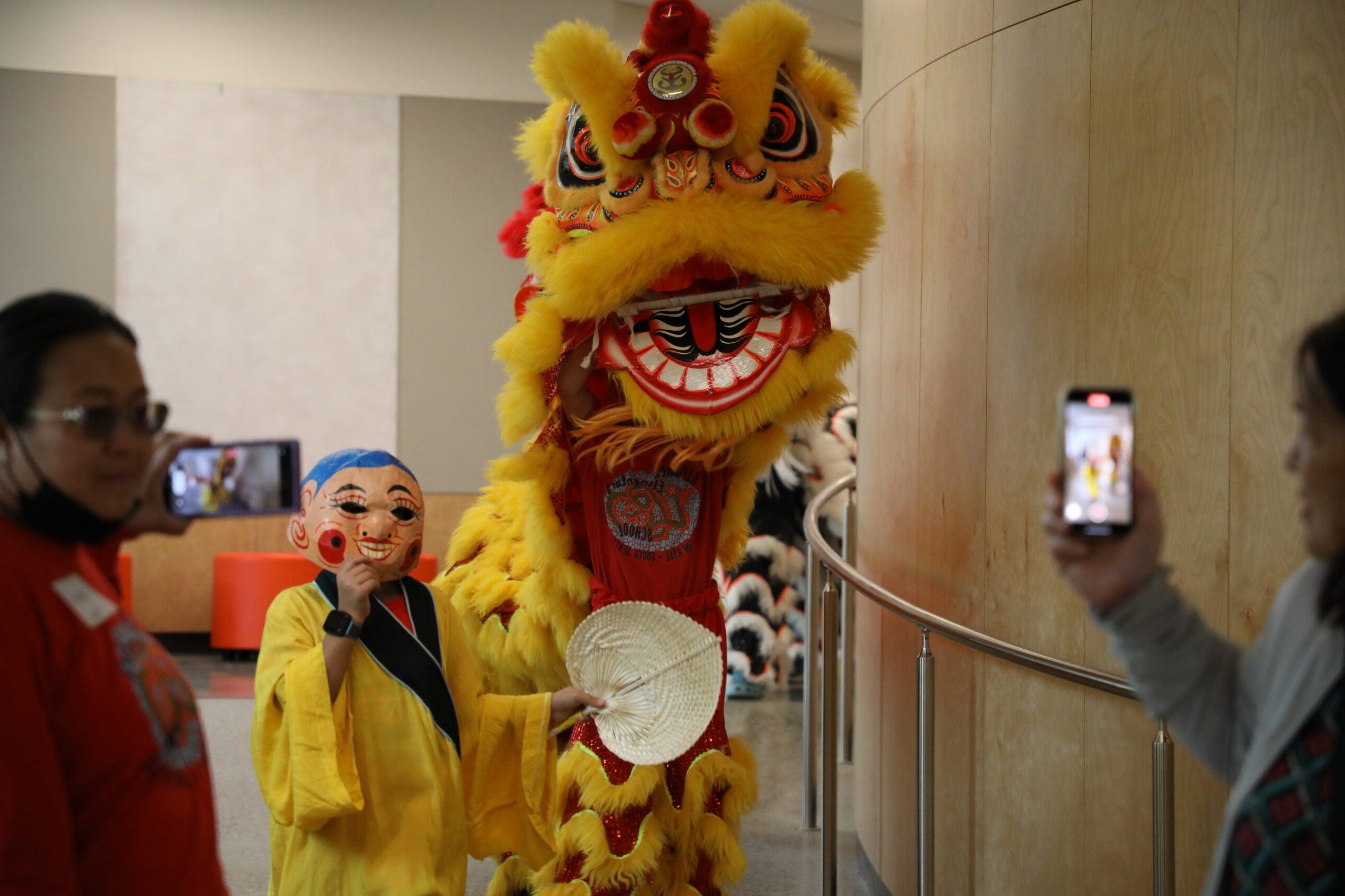 a yellow lion costume covers two people and a child holds a mask up next to it
